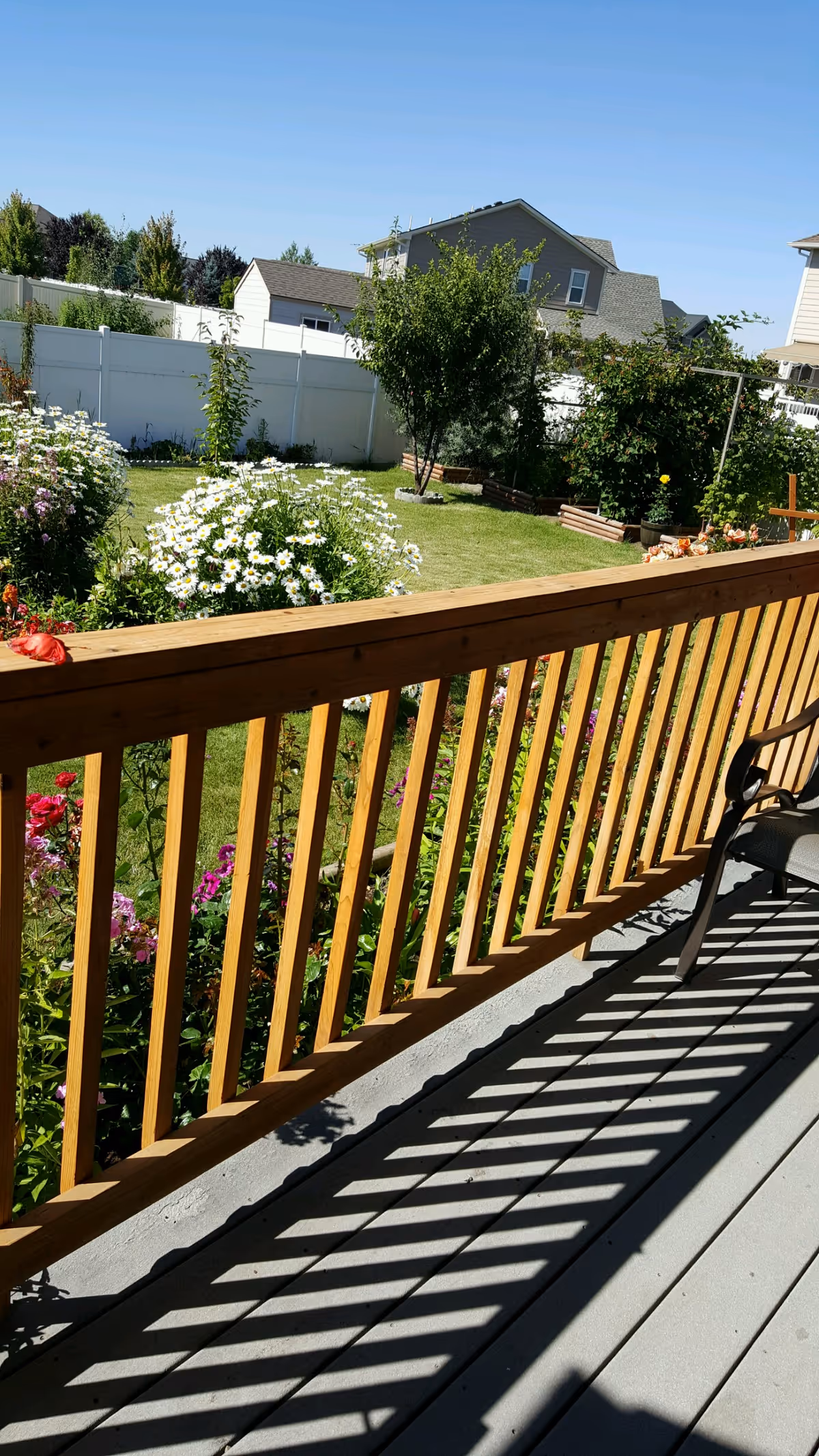 View from a wooden deck railing overlooking a garden with blooming flowers, green grass, and trees. In the background, there are houses and a white fence under a clear blue sky.