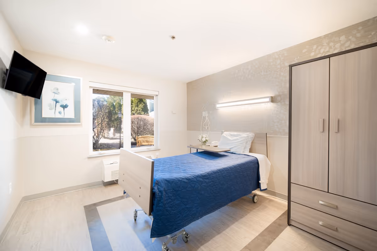A bright single-occupancy care room with a hospital-style bed dressed in a blue quilt, a wardrobe, wall-mounted TV, window looking outside, and a wall light.