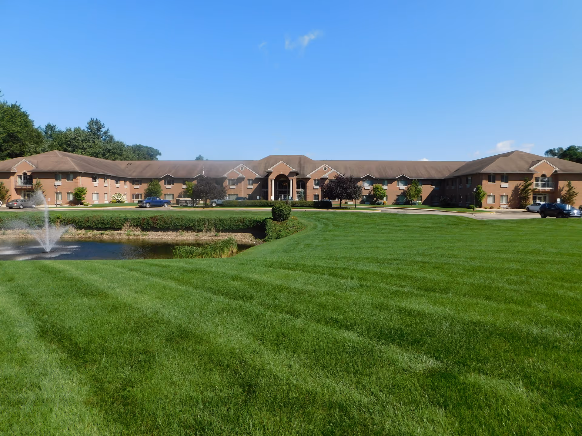 Wide view of Brentwood at Elkhart Independent Living facility showing a large, well-maintained green lawn with a small pond and fountain in the foreground. The two-story brick building with multiple windows and a central entrance is visible under a clear blue sky.