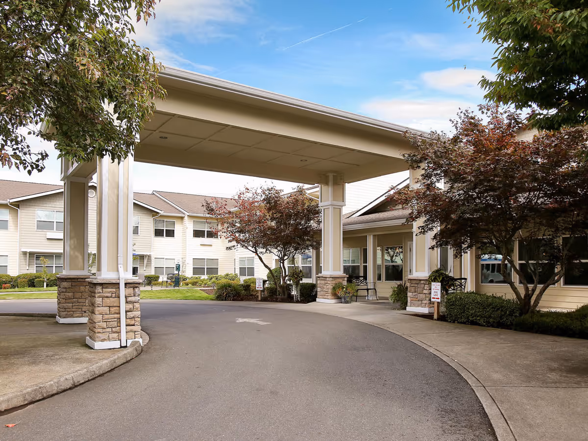 Covered porte-cochere and driveway leading to the front entrance of a multi-story senior living building with landscaped trees and shrubs.