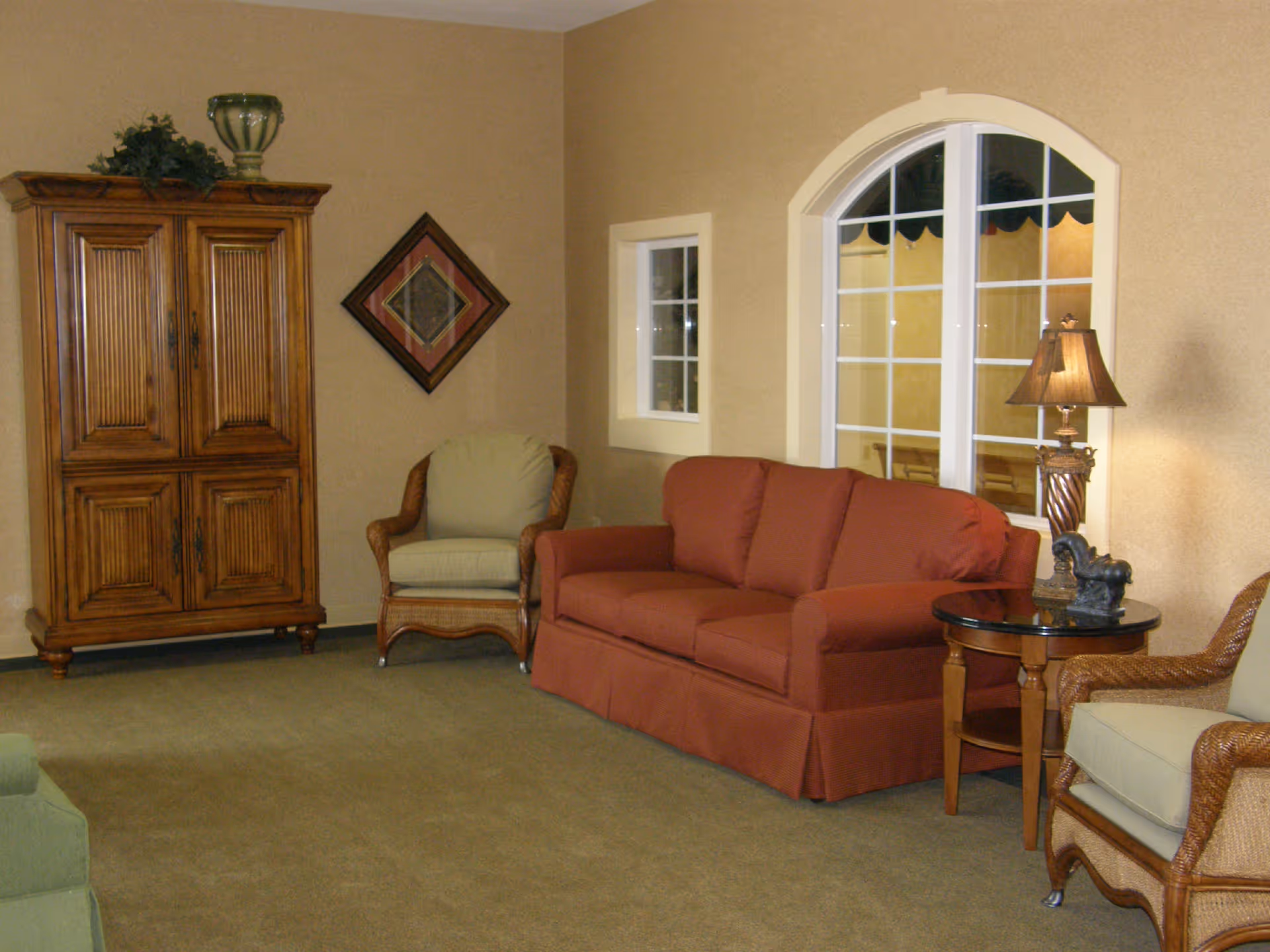 A cozy living room with a red sofa, two wicker armchairs with cushions, a wooden cabinet, a round side table with a decorative lamp and figurine, and a large arched window with white framing.