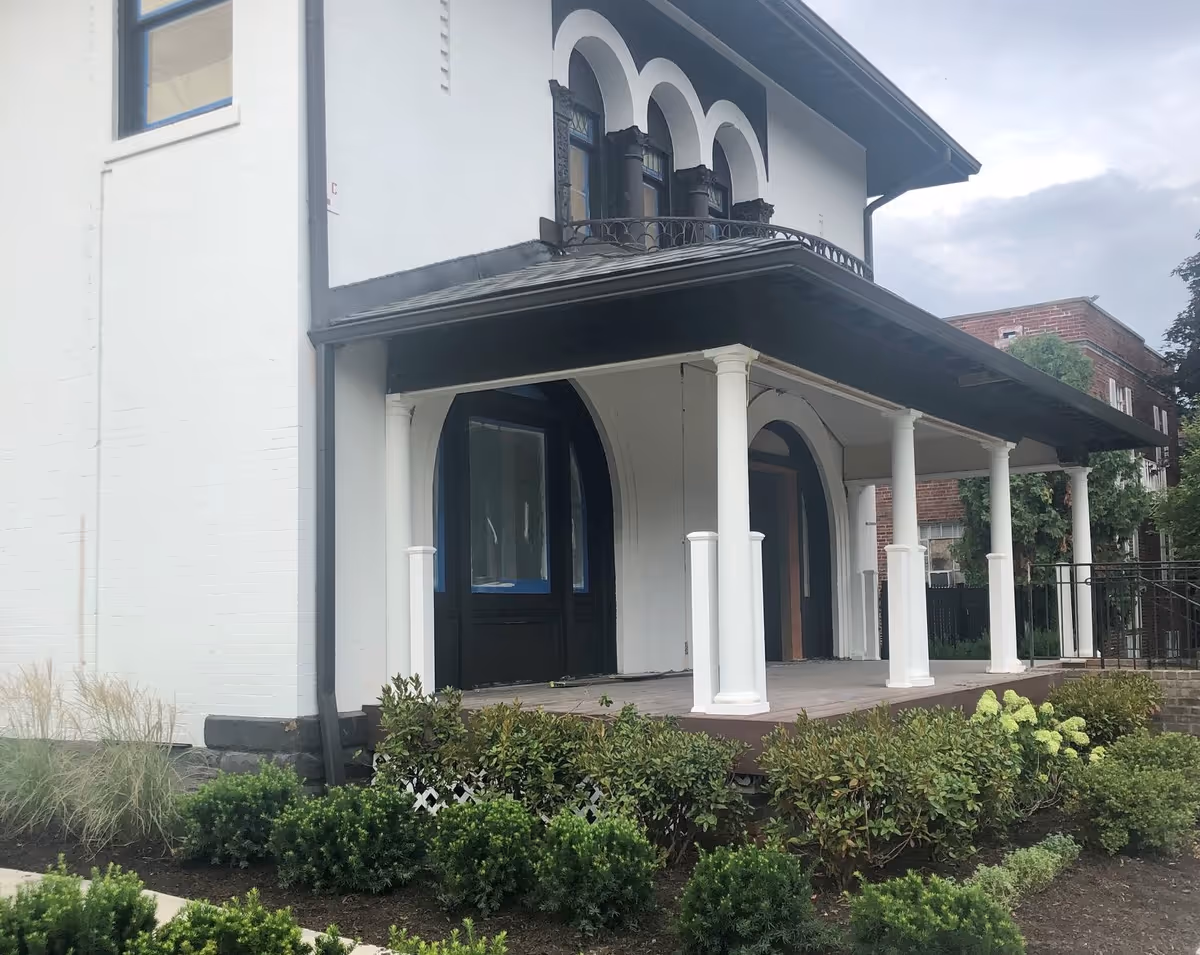 Front porch of a white two-story building with arched doorways, white columns, and landscaped shrubs.