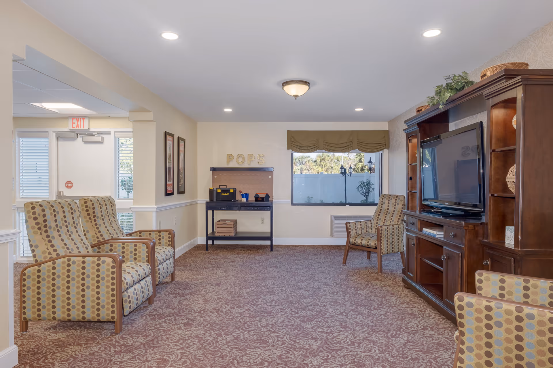 A common area in a senior living facility with patterned armchairs arranged around a large wooden entertainment center holding a flat-screen TV. The room has beige walls, a patterned carpet, and a window with a green valance. There is a black workbench with tools and the word 'POPS' on the wall above it. Exit doors and framed pictures are visible on the left side.