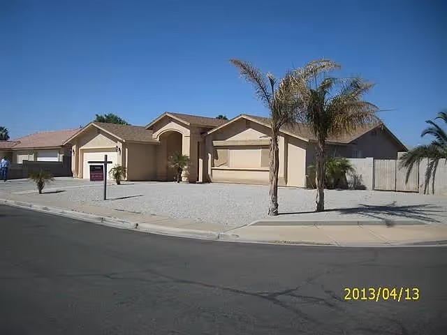 Single-story beige stucco house with a gravel front yard and a few palm trees, located on a corner lot under a clear blue sky.