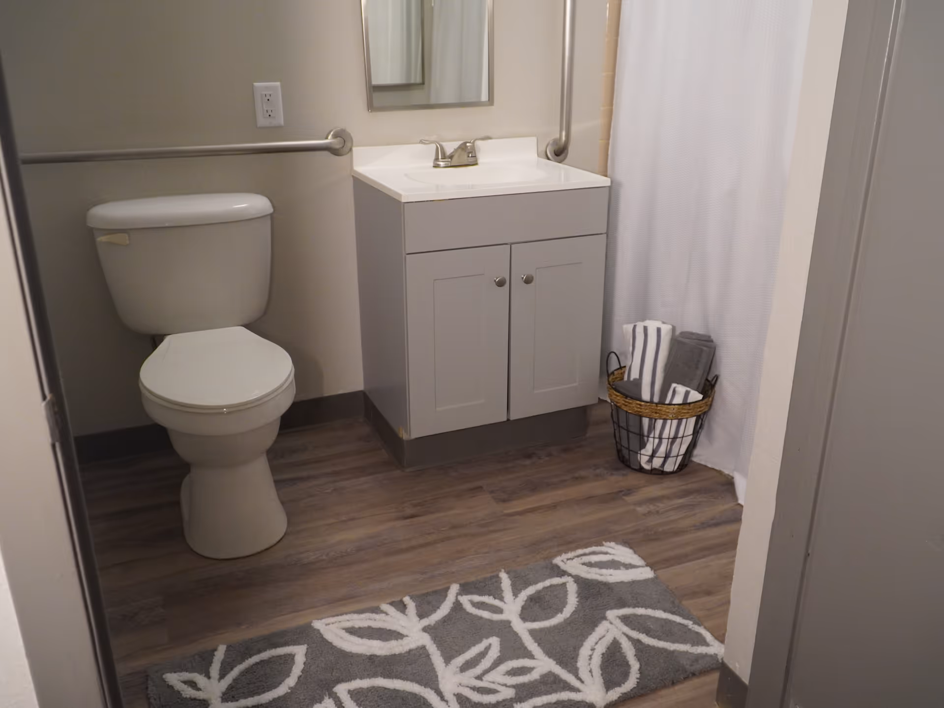 A clean bathroom with a white toilet, a white vanity with a sink and faucet, a mirror above the sink, a metal grab bar on the wall, a basket with folded towels, a white shower curtain, and a gray rug with a white leaf pattern on the wooden floor.