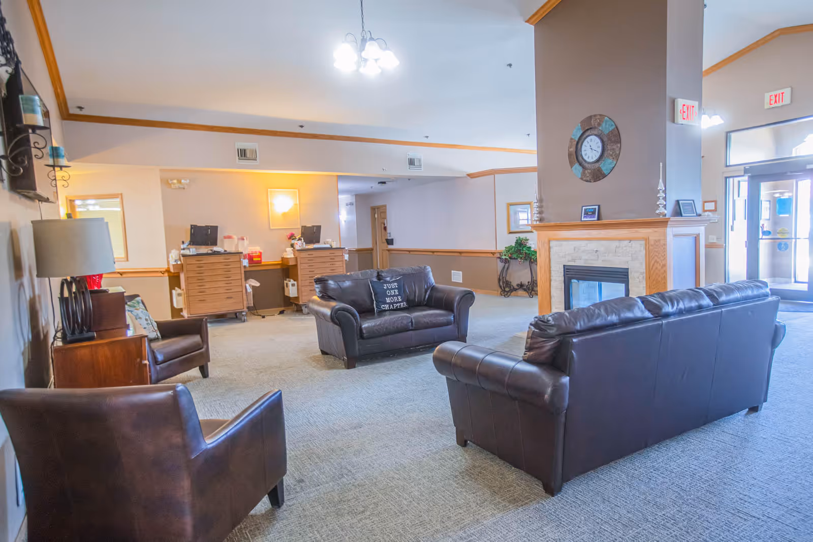 A bright and spacious senior living facility common area with leather sofas and armchairs arranged around a fireplace with a clock above it. The room has carpeted floors, light-colored walls with wooden trim, and a reception desk in the background. There are exit signs above the doors leading outside.
