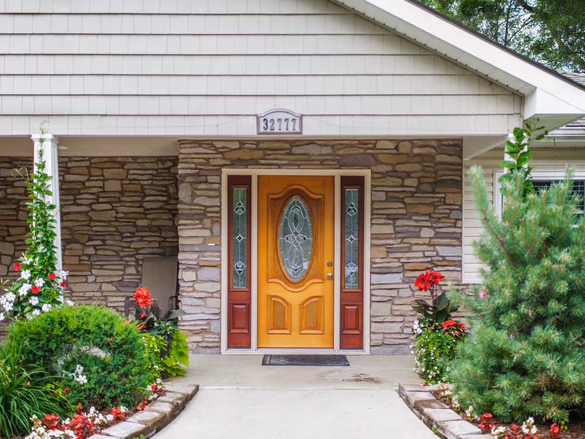 Front entrance of a senior care facility with a wooden door featuring decorative glass panels on either side. The exterior wall is made of stone, and there are well-maintained flower beds with various plants and flowers on both sides of the concrete walkway leading to the door.