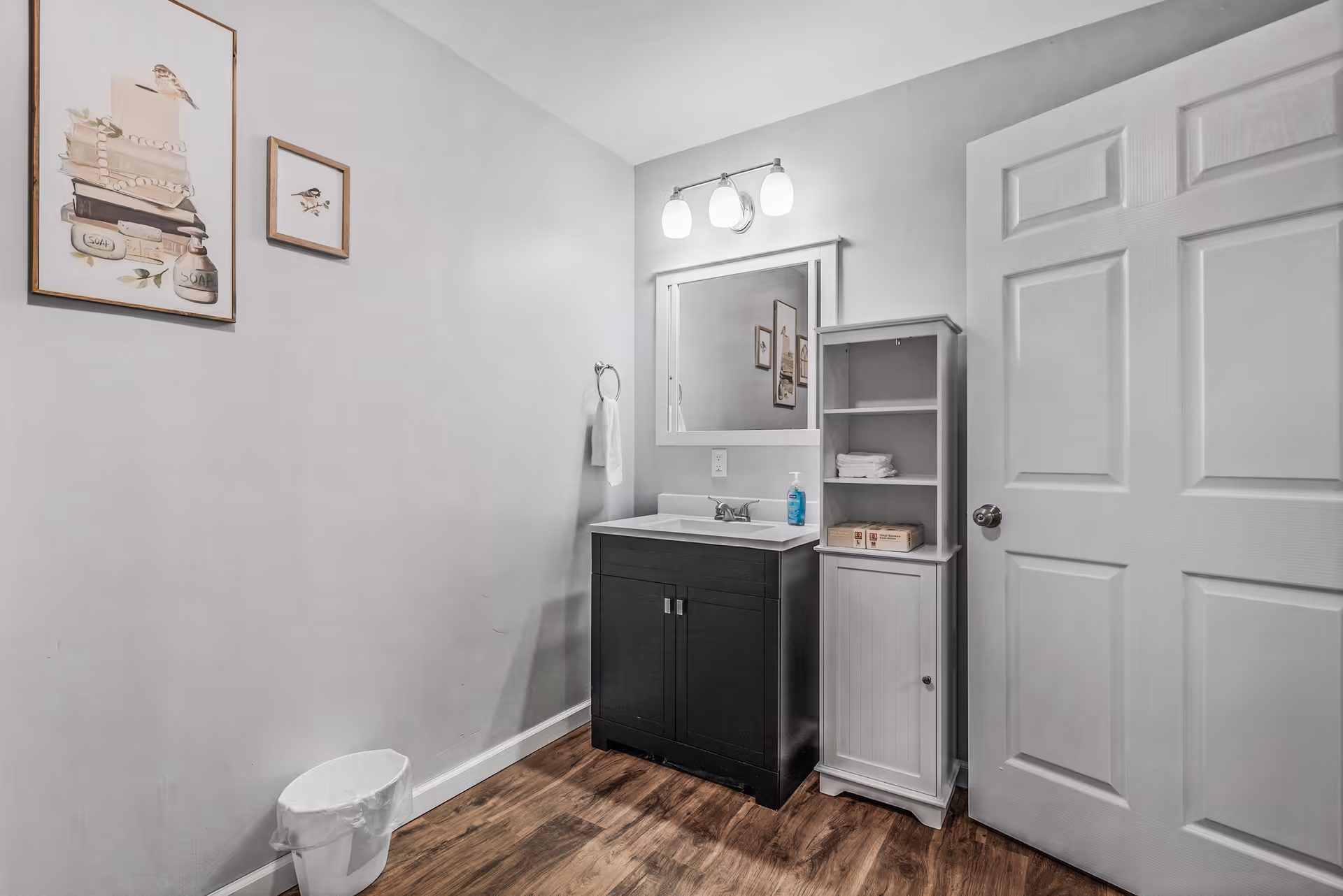 A clean bathroom with a dark wood vanity topped with a white sink and a mirror above it. There is a white cabinet with shelves next to the vanity holding folded towels and soap. The walls are light gray with two framed pictures of birds and books. The floor is wood, and a white door is visible on the right side.