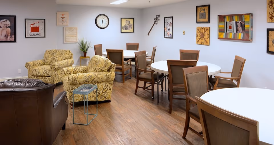 Community room with round dining tables and chairs and a seating area with upholstered armchairs against a wall with framed artwork.