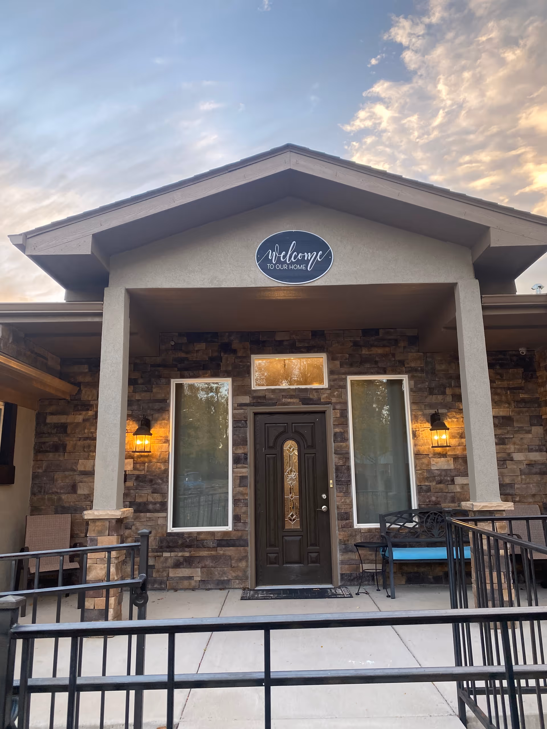 Front entrance of a senior living facility with a covered porch, stone facade, two tall windows on either side of a dark wooden door with decorative glass, outdoor wall lanterns lit on both sides, a bench with blue cushion, and a sign above the door that reads 'welcome TO OUR HOME'.