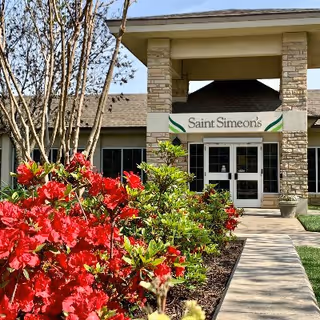 Entrance of Saint Simeon's Senior Community building with a covered porch supported by stone pillars, a sign displaying the facility's name above the double glass doors, and vibrant red flowers and green shrubs lining the walkway leading to the entrance.