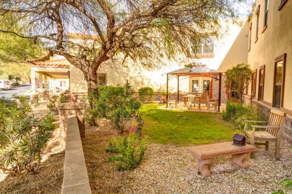 Outdoor garden area at Tucson Place at Ventana Canyon featuring a small grassy lawn, a tree, shrubs, a stone bench, wooden chairs, and a gazebo with a table and chairs under it. The building walls and windows are visible in the background.