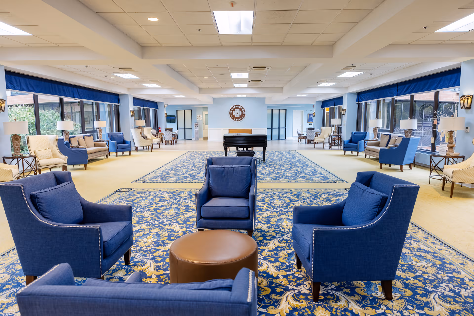 Spacious senior living facility common area with blue and beige armchairs arranged around a round brown ottoman on a patterned blue and yellow carpet. Large windows with blue valances line both sides of the room, allowing natural light to fill the space. A black grand piano is positioned in the center near the back wall, which has a circular sign reading 'Westminster Canterbury'. Additional seating areas with sofas and chairs are visible along the sides of the room.