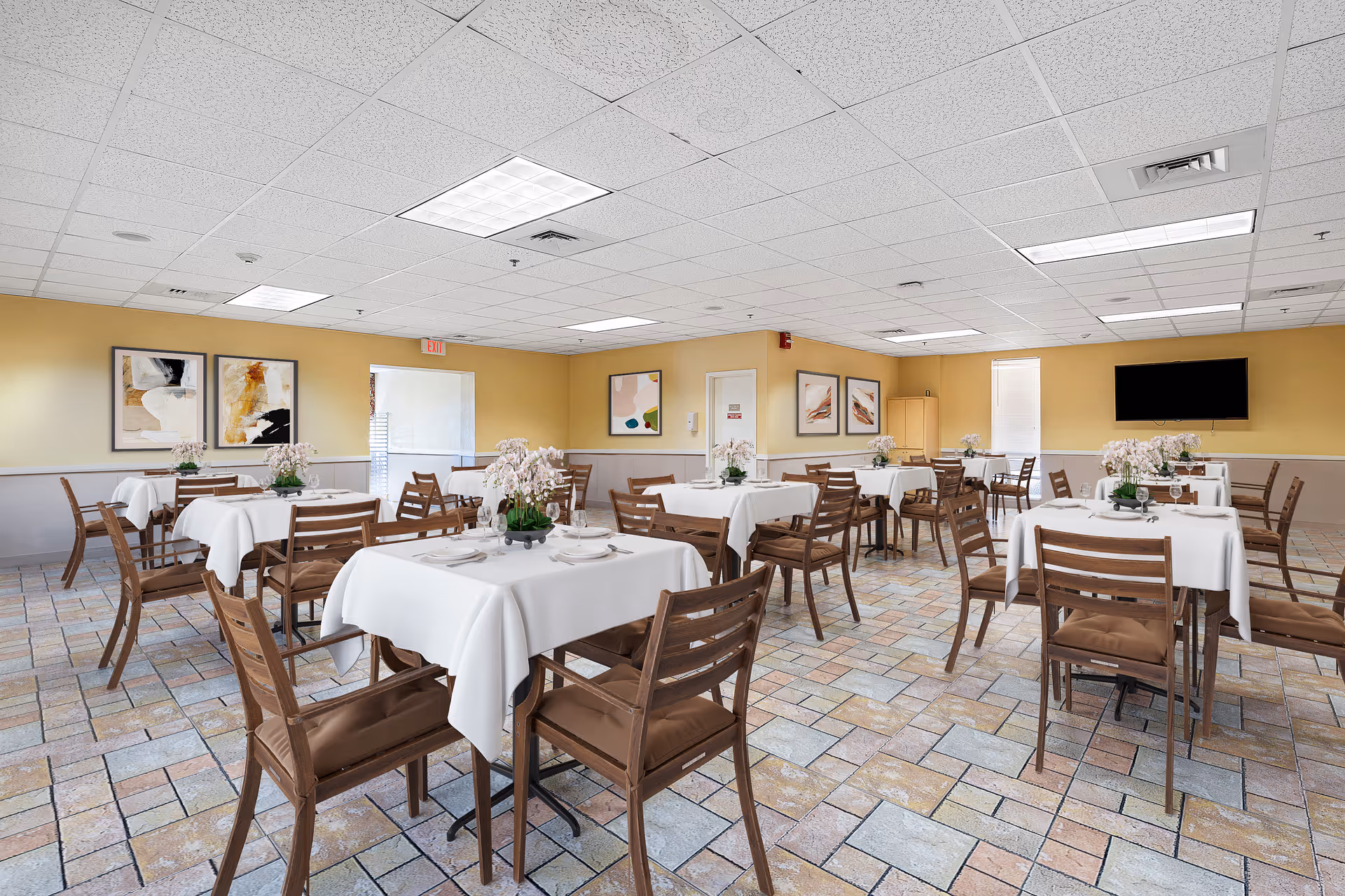 Bright dining room with multiple white-clothed tables, wooden chairs, floral centerpieces, and a wall-mounted TV.