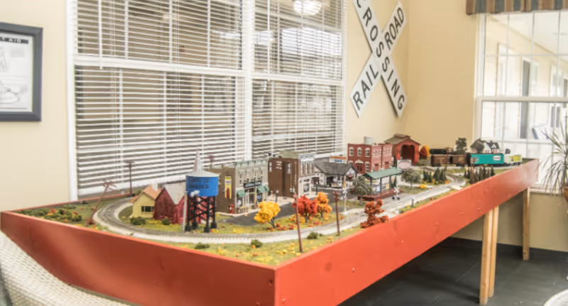 A detailed model train set displayed on a large red table inside a room with windows and blinds. The model includes miniature buildings, trees with autumn colors, a water tower labeled 'Town of Greece,' and a railroad crossing sign mounted on the wall behind the table.