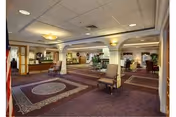 Spacious interior common area of a senior living facility with carpeted floors, multiple seating areas with chairs, decorative columns, and warm lighting fixtures on the ceiling and walls.