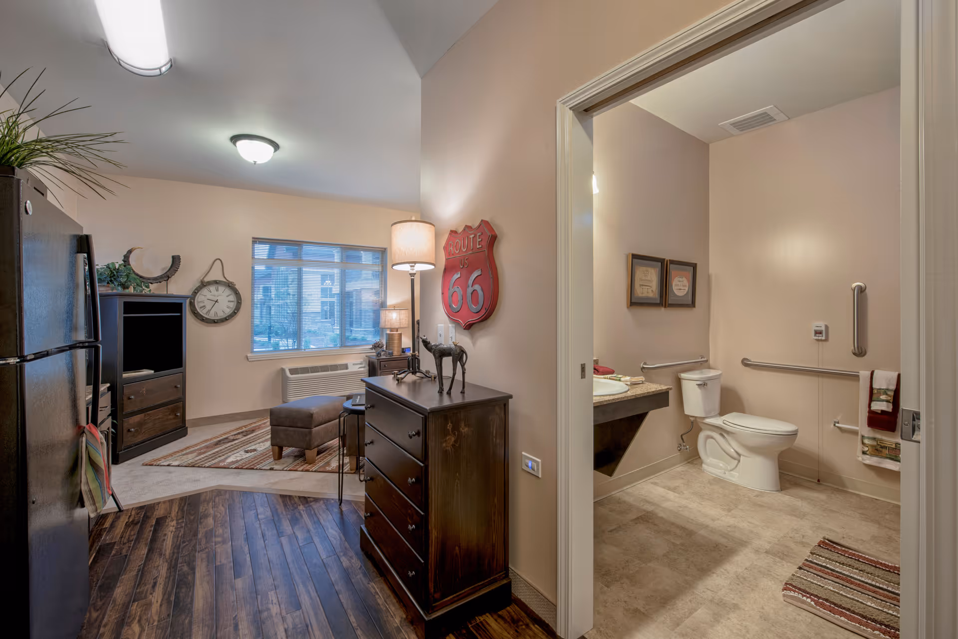 Interior view of a senior living facility room showing a kitchenette area with a refrigerator, a wooden dresser with a lamp and decorative items, a window with blinds, and a bathroom with a toilet, sink, grab bars, and framed pictures on the wall.
