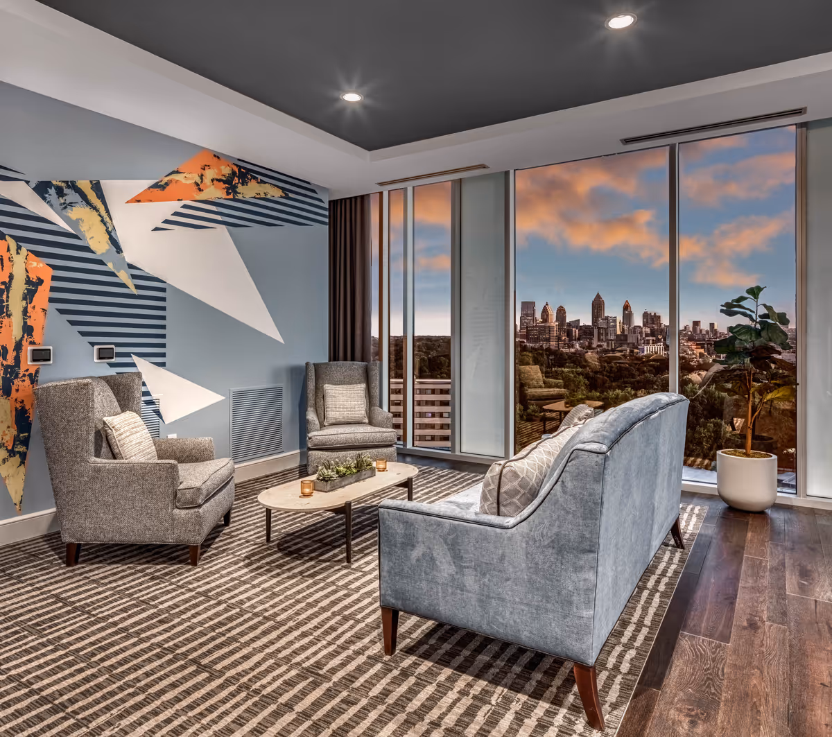 Modern senior living lounge area with two gray armchairs, a gray sofa, a small coffee table with candles and a plant centerpiece, a patterned carpet, and large floor-to-ceiling windows showing a city skyline at sunset. The wall features an abstract geometric mural with blue, white, and orange shapes.