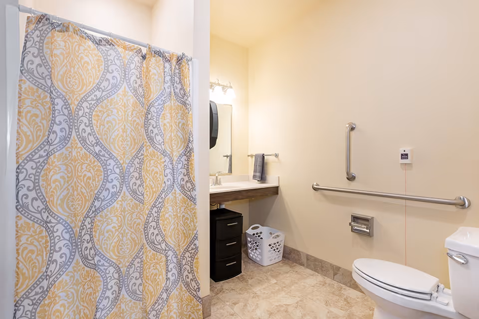 A bathroom with a patterned yellow and gray shower curtain, a white toilet with grab bars on the wall, a sink with a mirror and light fixture above it, a black storage drawer unit, and a white laundry basket on a tiled floor.