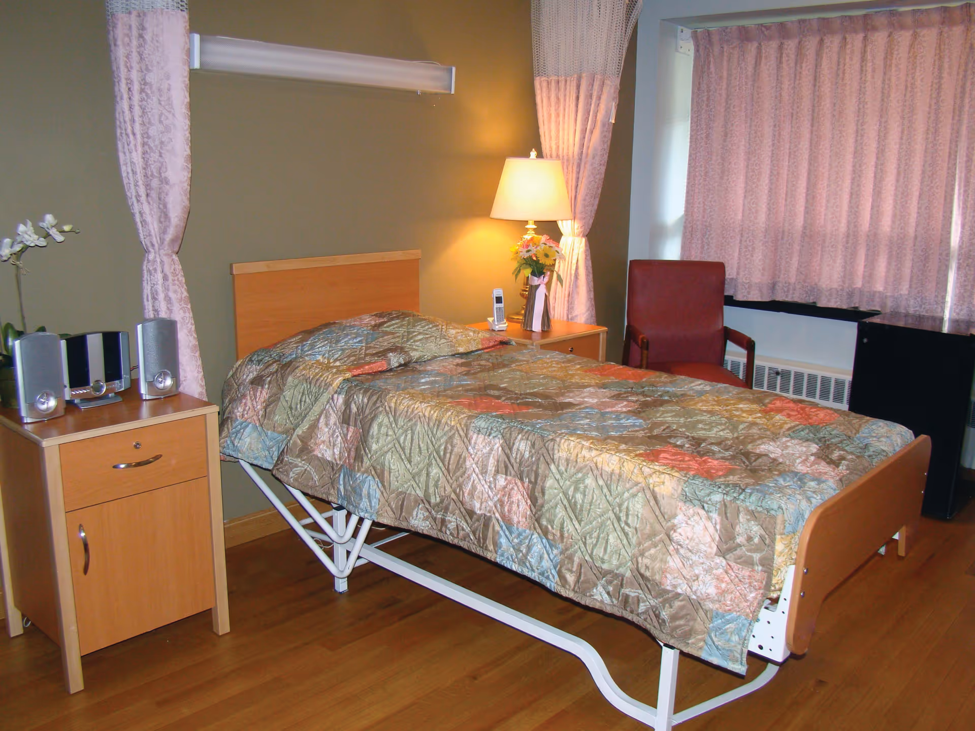A single bed with a colorful quilted bedspread in a room with wooden flooring. Next to the bed is a wooden nightstand with a stereo system and a white orchid plant. On the other side of the bed is another nightstand with a lamp, a vase of flowers, and a cordless phone. A red armchair is positioned near a window with pink curtains.