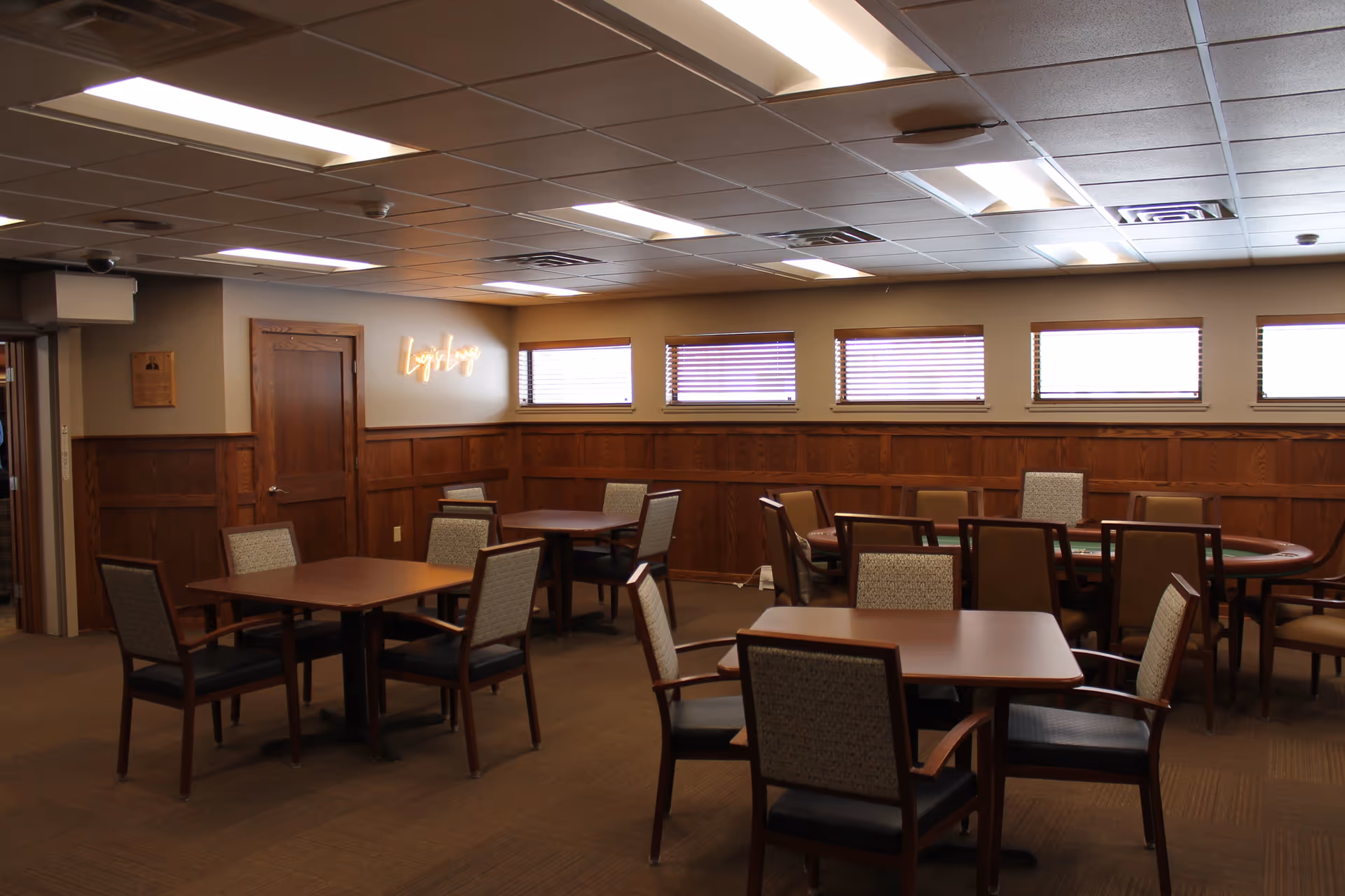 Spacious communal dining/activity room with multiple tables and chairs, wood-paneled walls, and small windows near the ceiling.