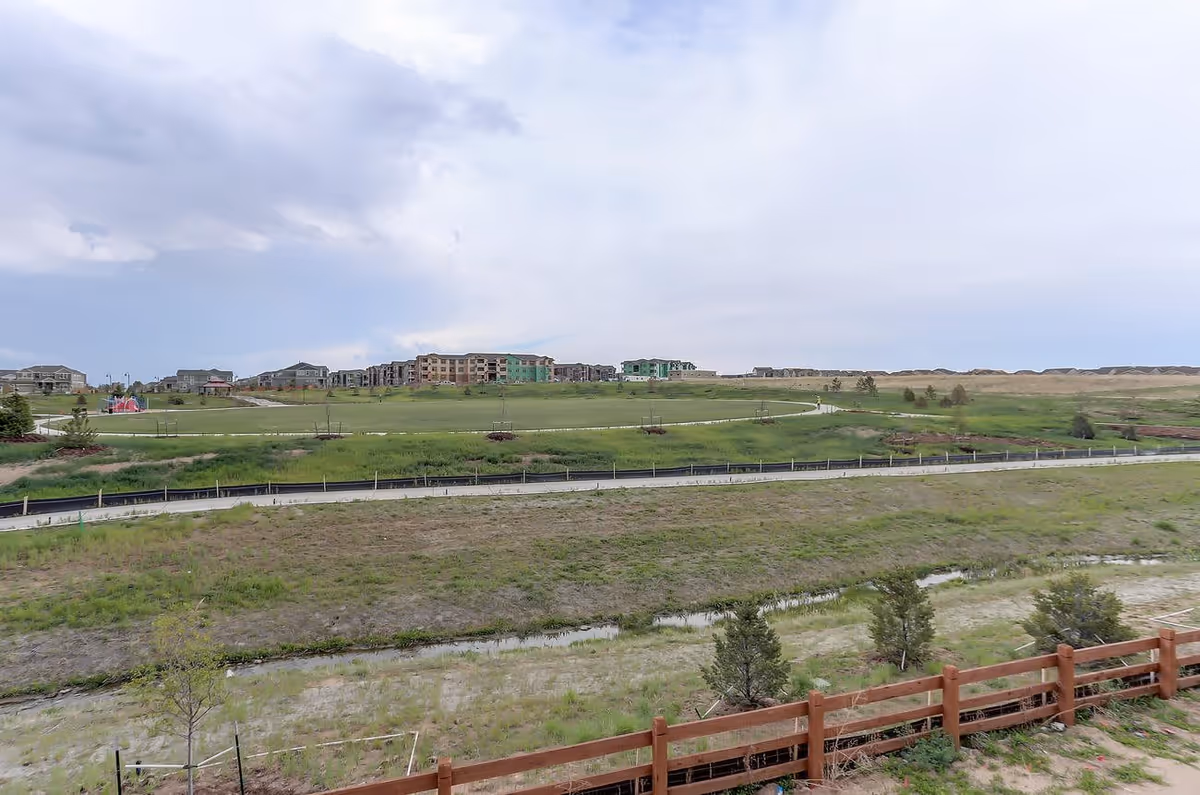 A wide view of a grassy open field with a paved walking path running through it. In the distance, there are several multi-story residential buildings under a partly cloudy sky. A wooden fence and some small trees are visible in the foreground.