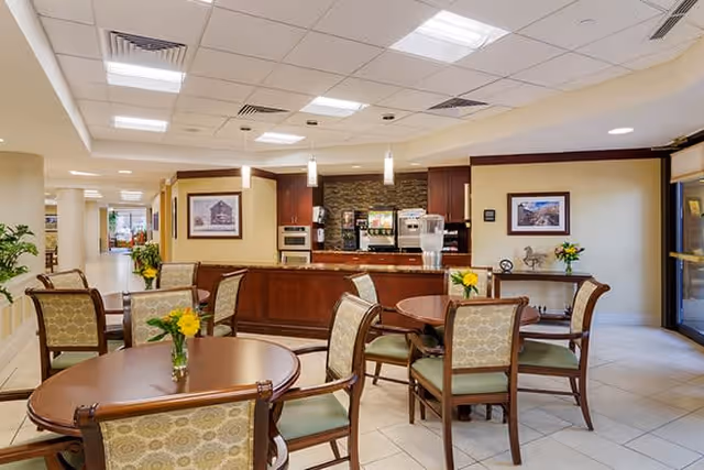 A well-lit common area with round wooden tables and cushioned chairs arranged neatly. Each table has a small vase with yellow flowers. In the background, there is a counter with a coffee machine and water dispenser, framed pictures on the walls, and a hallway leading to other parts of the facility.
