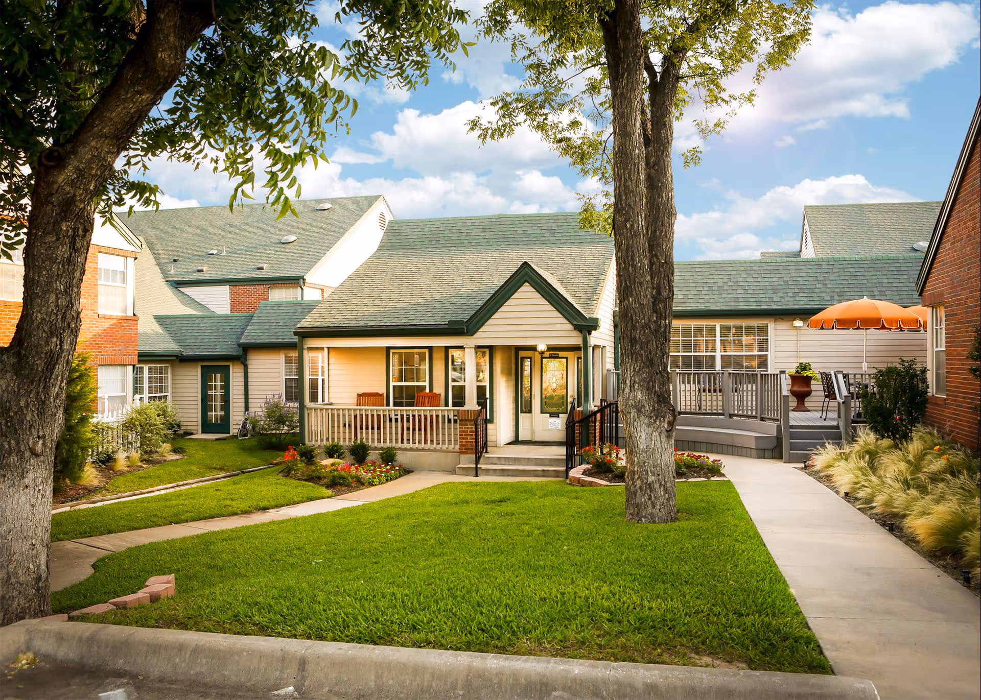 Exterior view of Bethesda Gardens Arlington Assisted Living facility showing a well-maintained lawn, trees, flower beds, a paved walkway, and a building with green roofs and a porch with chairs. There is also a deck area with an orange umbrella and outdoor seating.
