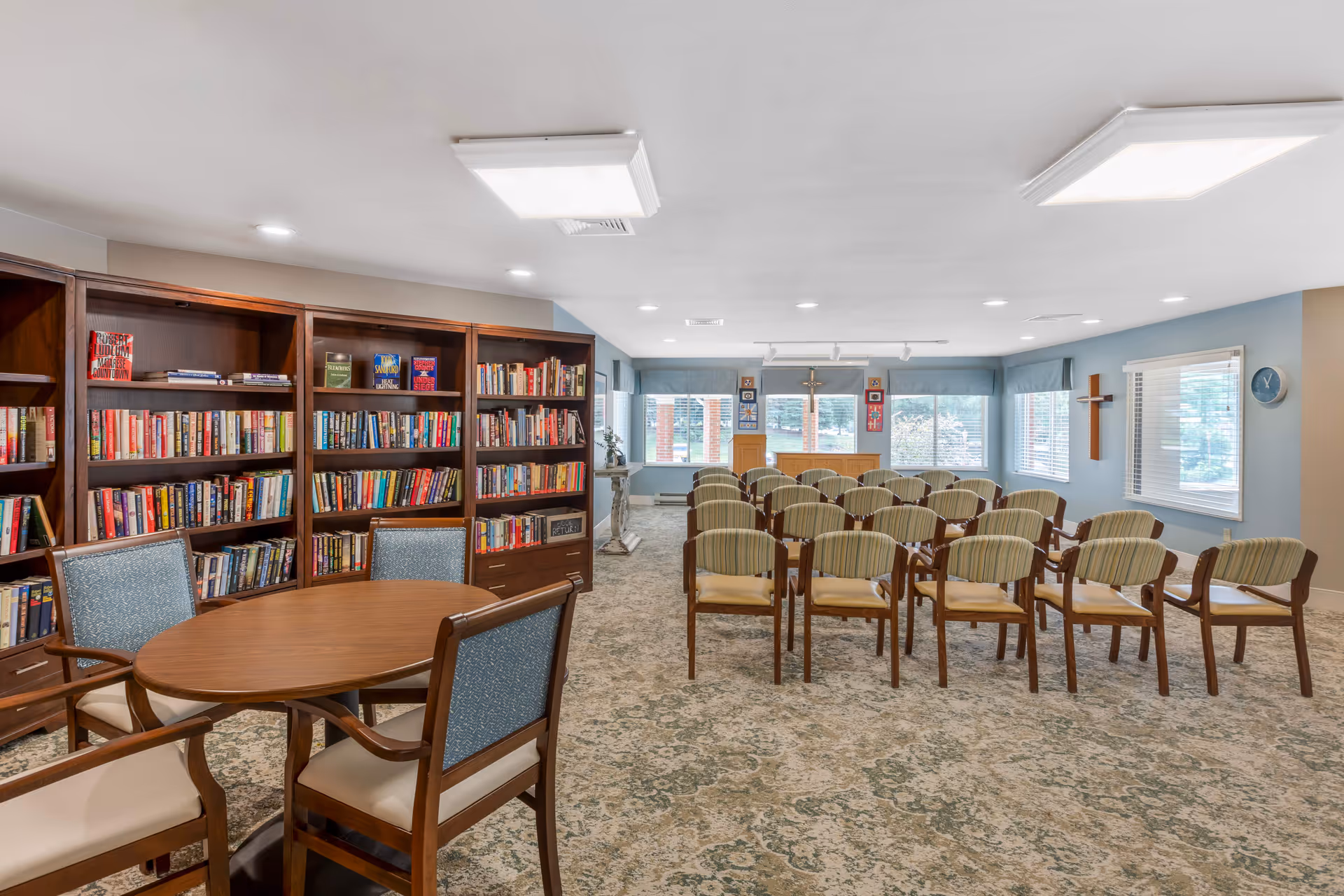 A bright senior living community room with bookshelves and a round table on the left and rows of chairs facing a podium and windows.