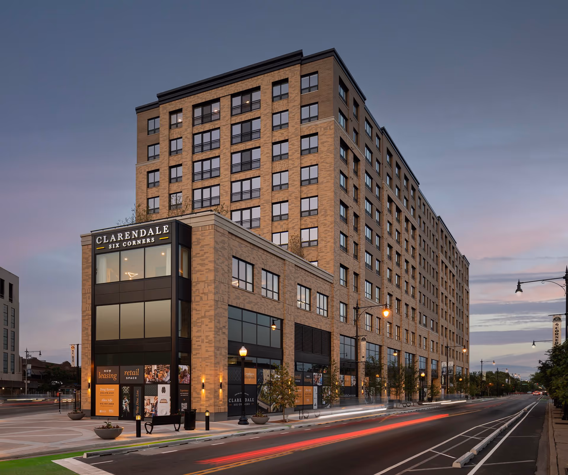 Exterior view of Clarendale Six Corners, a multi-story brick building with large windows and retail space on the ground floor, situated along a street with streetlights and some greenery at dusk.