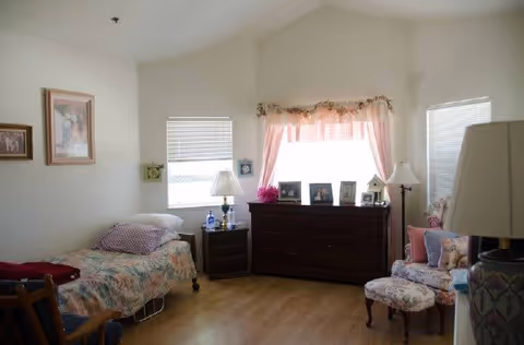 Sunlit bedroom with a single bed, wooden dresser beneath a curtained window, and an upholstered chair with ottoman.
