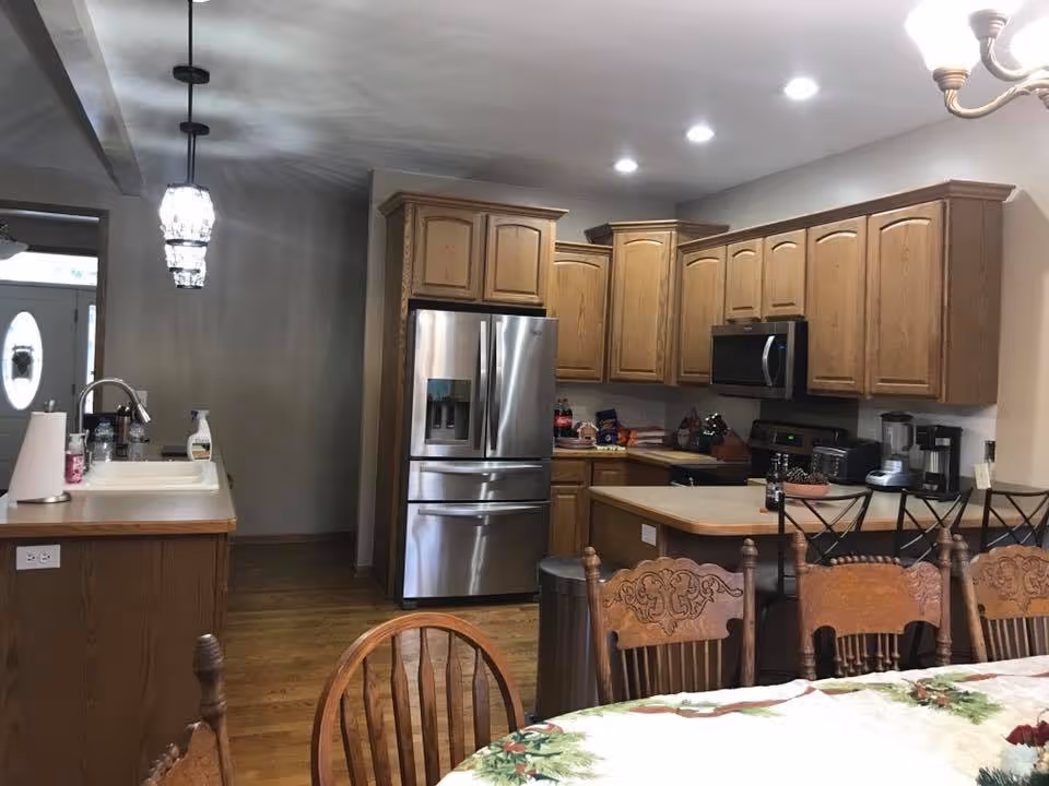Interior view of a kitchen with wooden cabinets, a stainless steel refrigerator, microwave, and various kitchen appliances on the counters. A dining table with a floral tablecloth and wooden chairs is visible in the foreground. The kitchen has hardwood floors and pendant lighting above the sink area.