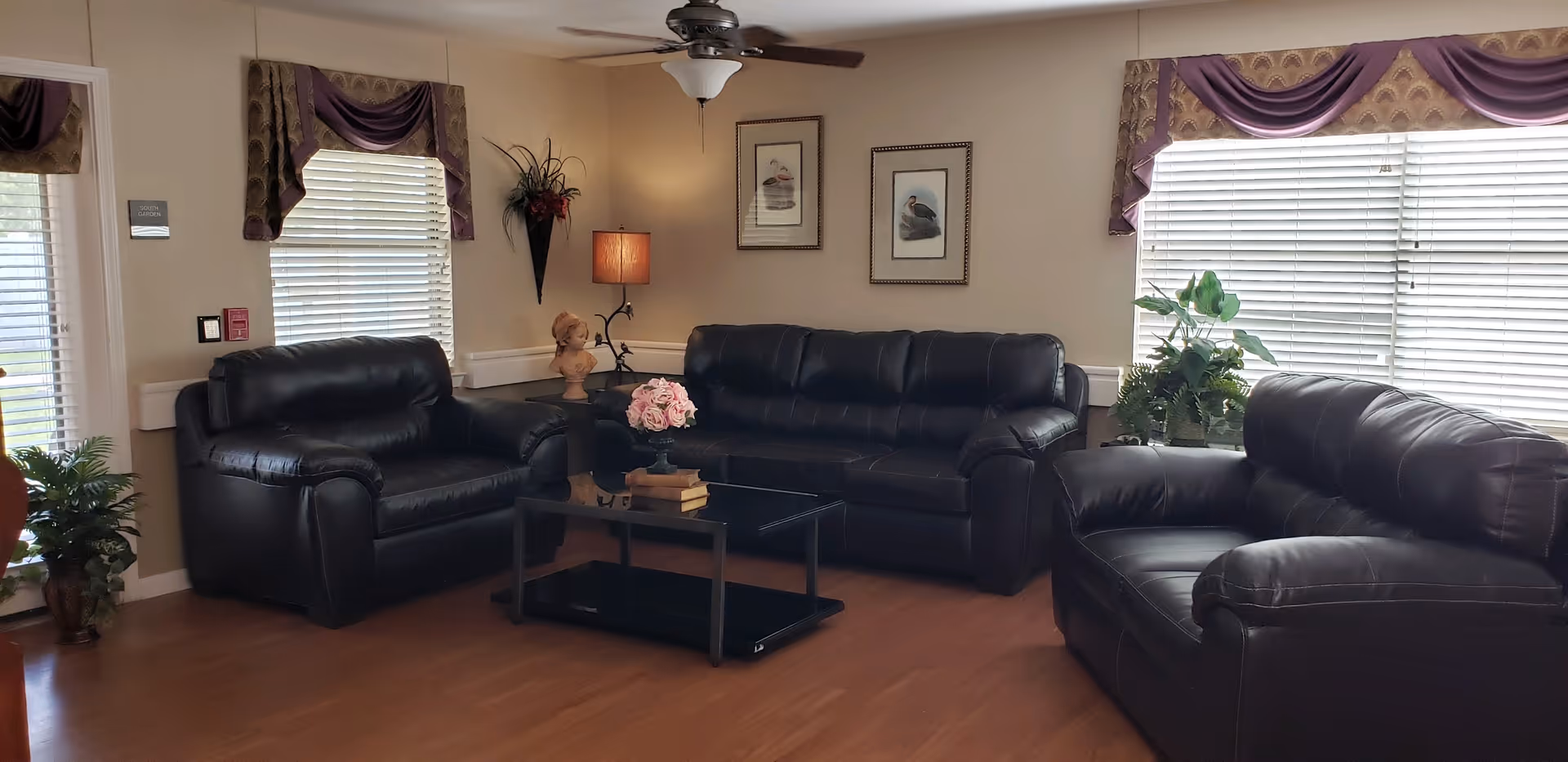 Comfortable lounge with dark leather sofas around a coffee table, plants, lamps, and large windows with valances.