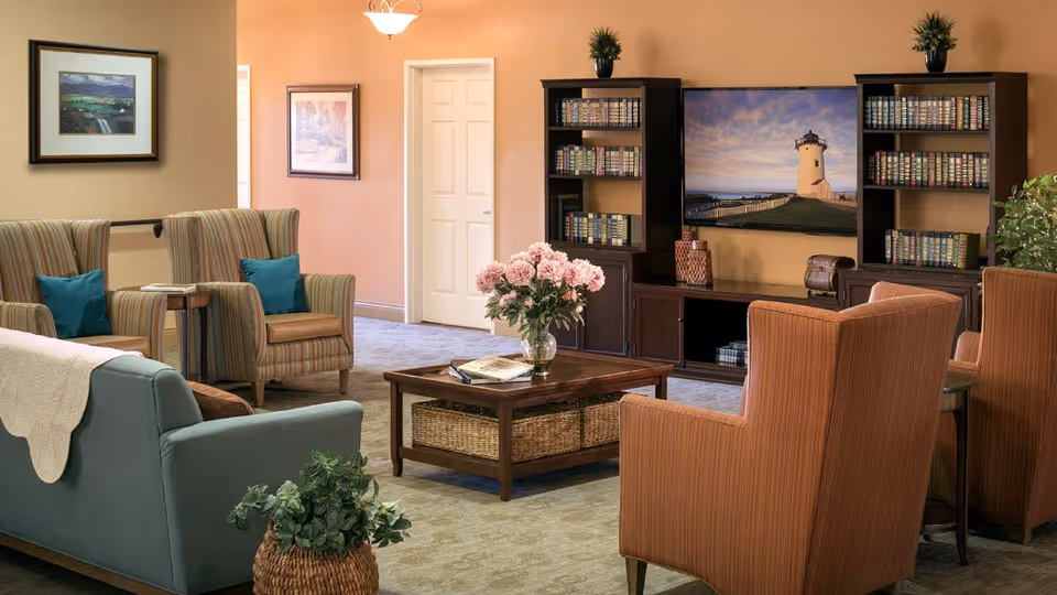 A cozy living room with a blue sofa and four striped armchairs arranged around a wooden coffee table with a vase of pink flowers. Behind the seating area, there are two dark wooden bookshelves filled with books and a large flat-screen TV displaying an image of a lighthouse. The walls are painted in warm tones with framed artwork, and there are plants adding greenery to the space.