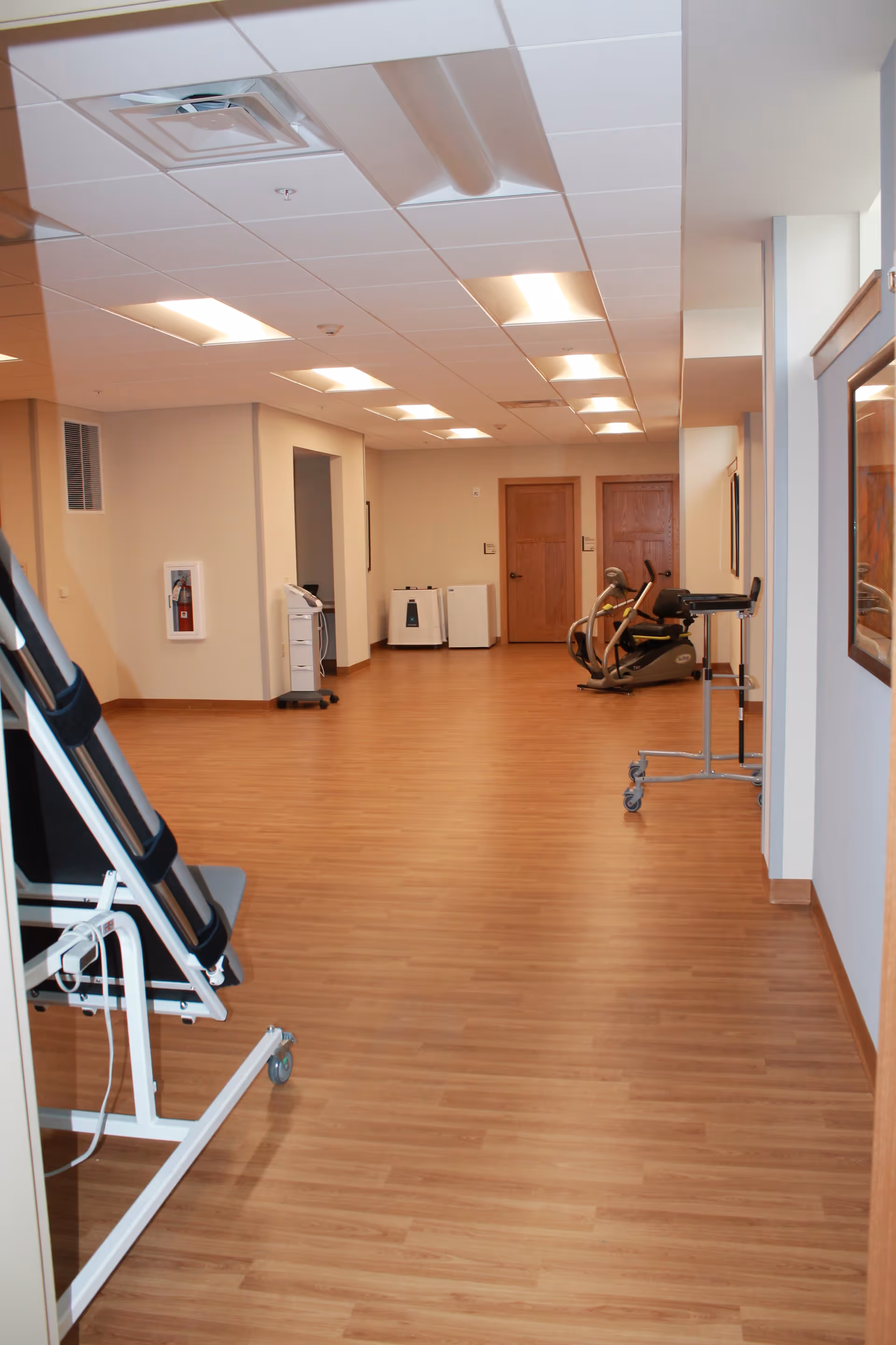 Interior hallway of a senior living facility with wood flooring, beige walls, ceiling lights, and exercise equipment including a recumbent bike and a standing frame. Two wooden doors are visible at the end of the hallway.