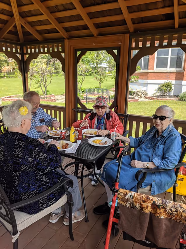 Four elderly individuals sitting around a black metal table under a wooden gazebo, enjoying a meal together. The setting is outdoors with green grass and trees visible in the background. One person is wearing a red jacket and a floral hat, another is in a blue denim outfit with sunglasses, and the others are dressed casually. There are plates of food, condiments, and drinks on the table.