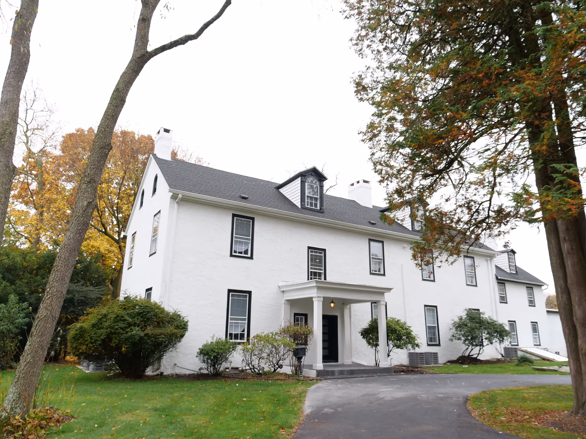 Exterior view of a large white two-story building with black-trimmed windows, a small covered entrance, surrounded by green grass, bushes, and tall trees with autumn foliage.