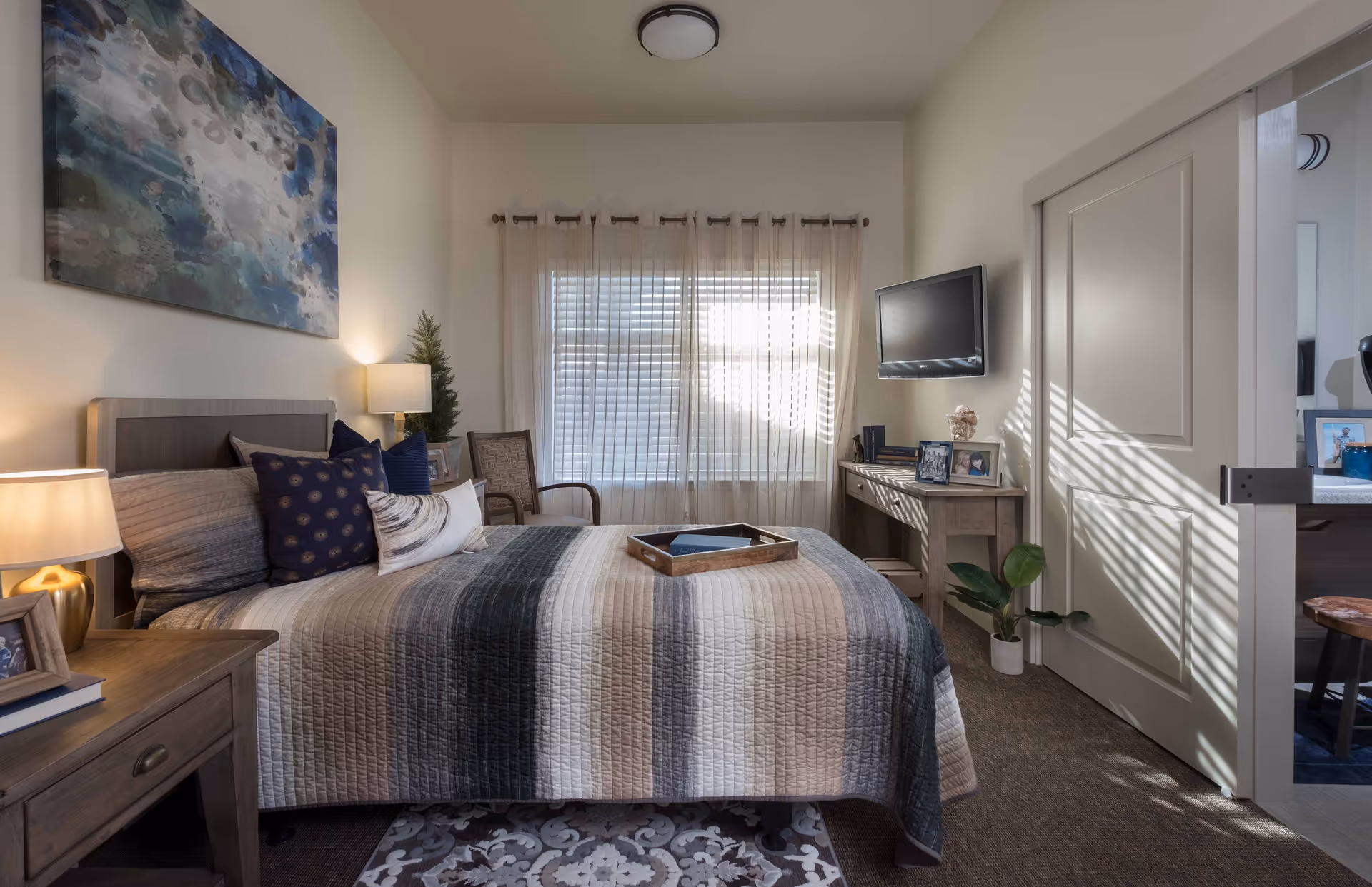 A cozy bedroom with a neatly made bed featuring a striped quilt and multiple pillows. There is a wooden nightstand with a lamp and framed photos on the left side. A window with sheer curtains lets in natural light, and a flat-screen TV is mounted on the wall opposite the bed. A small desk with framed photos and decorative items is positioned under the TV. The room has neutral walls, a patterned rug, and a door leading to another room.