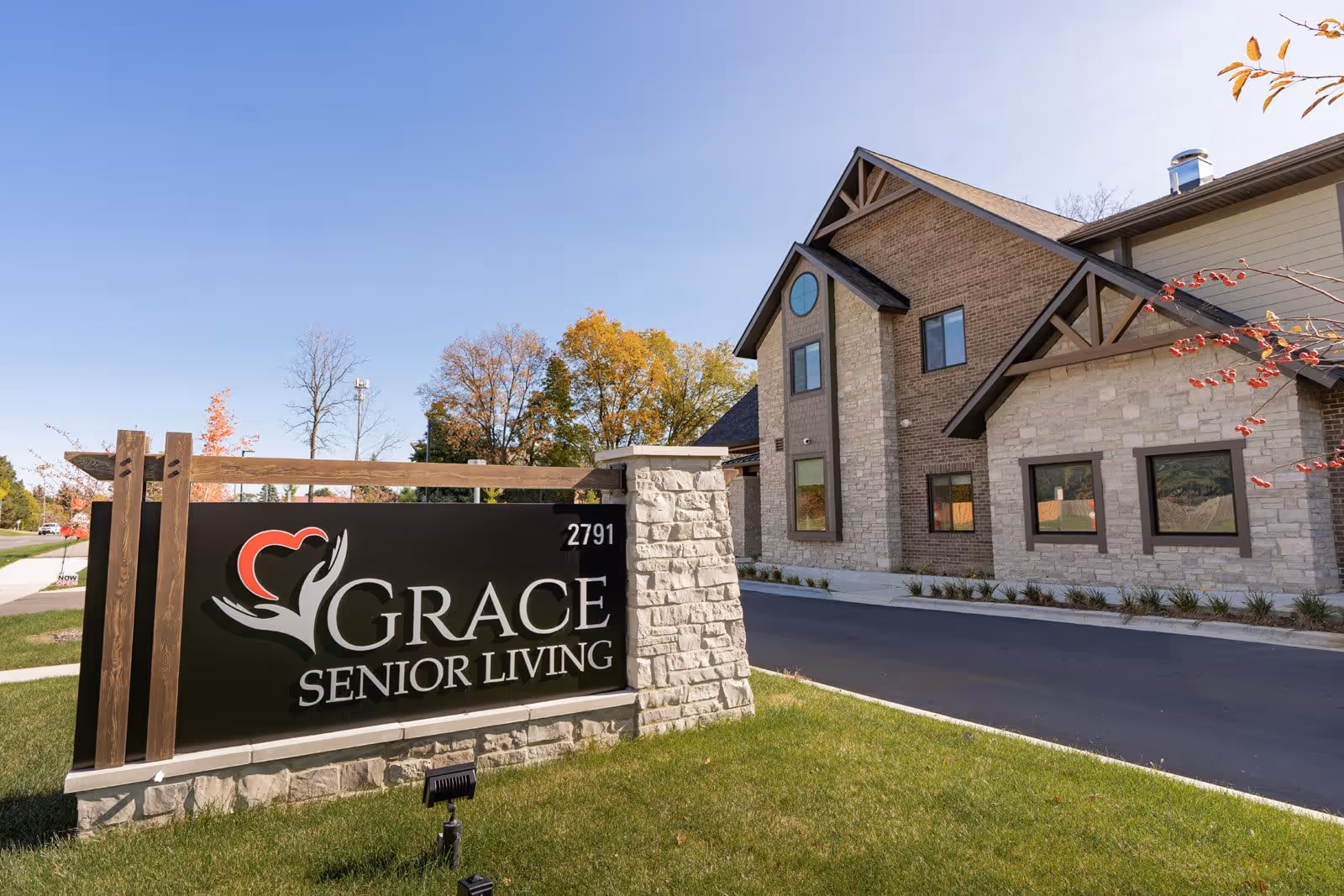 Exterior view of Grace Senior Living Rochester Hills building with a large sign in the foreground displaying the facility's name and logo. The building features stone and brick walls with multiple windows and a peaked roof. The sky is clear and blue, and there is green grass and some trees with autumn foliage around the property.