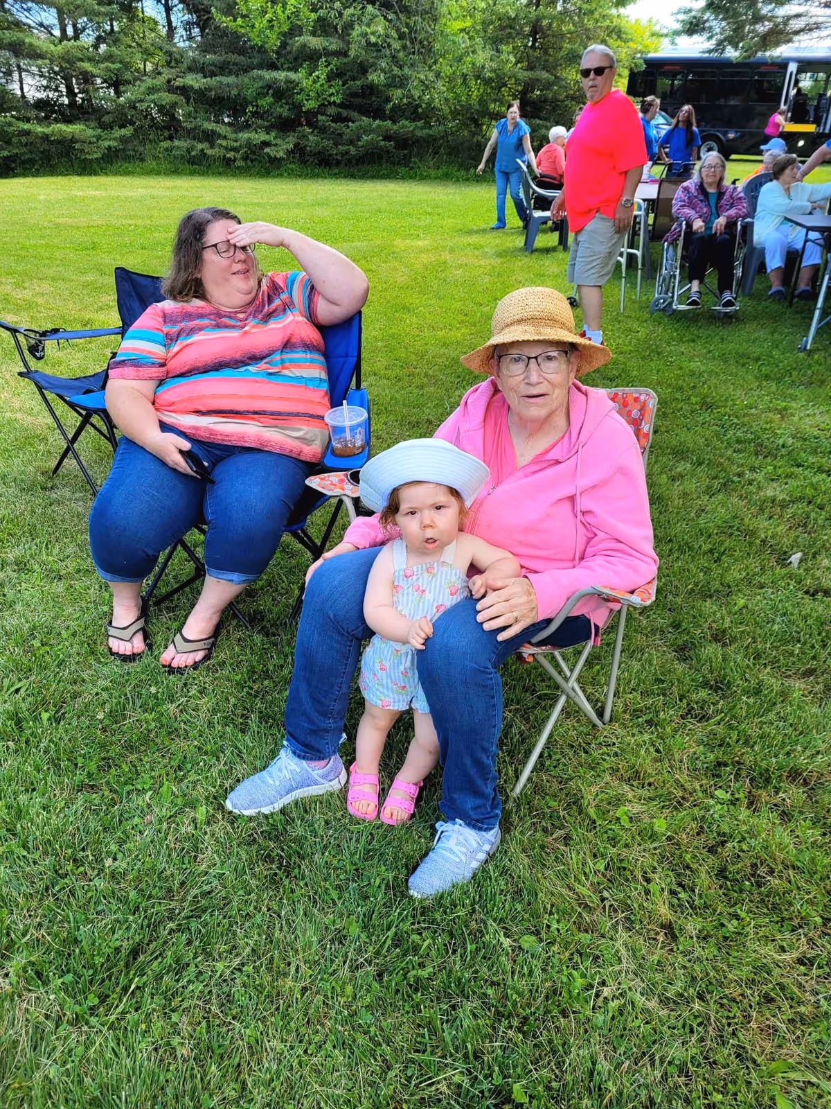 A group of people gathered outdoors on a grassy area. In the foreground, an elderly woman wearing a straw hat and pink jacket sits in a folding chair with a young child in a white hat and floral outfit sitting on her lap. Next to them, a woman in a striped shirt and jeans sits in another folding chair holding a drink. In the background, several other people, including an elderly person in a wheelchair and a man in a bright pink shirt, are visible near tables and a bus, with trees and greenery surrounding the area.