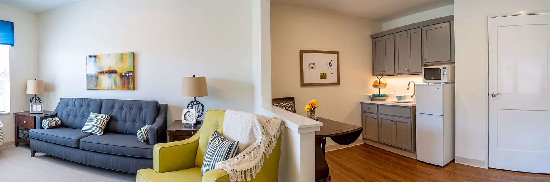 Bright furnished living area with a gray sofa, green armchair, side tables and lamps, and a small kitchenette with gray cabinets and a fridge.