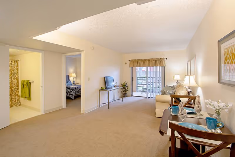 Spacious living room of an assisted living apartment with a sofa, TV, small dining table, and sliding glass door to a balcony, with bedroom and bathroom doorways visible.