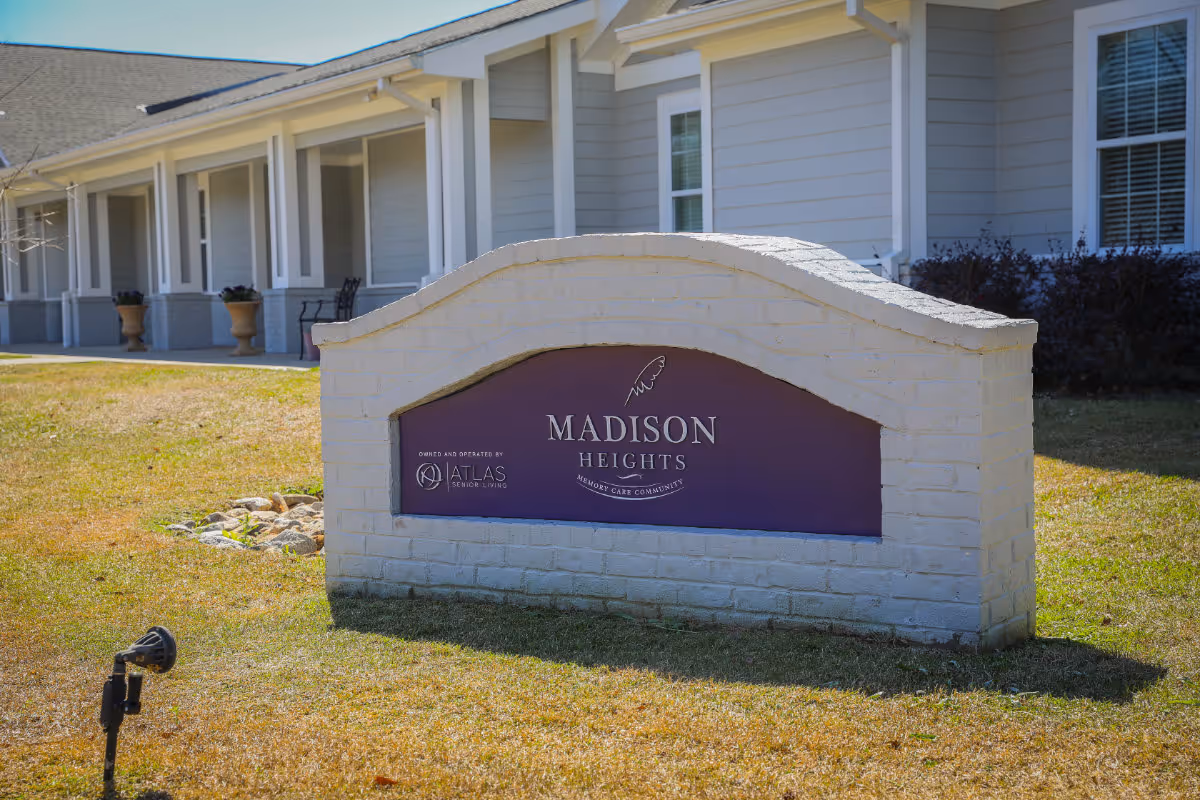 A white brick sign in front of a building with the text 'Madison Heights Memory Care Community' and 'Owned and operated by Atlas Senior Living'. The building behind the sign has a light gray exterior with white trim and several windows and columns.