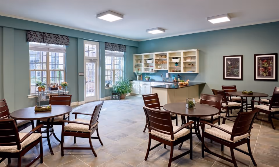 A bright and spacious common area with several round tables and cushioned wooden chairs arranged around them. The room features large windows with floral valances, letting in natural light. There is a kitchenette with white cabinets, a countertop, and open shelves decorated with plants and small items. The walls are painted a soft green, and two framed artworks hang on one wall. The floor is tiled in a neutral color.