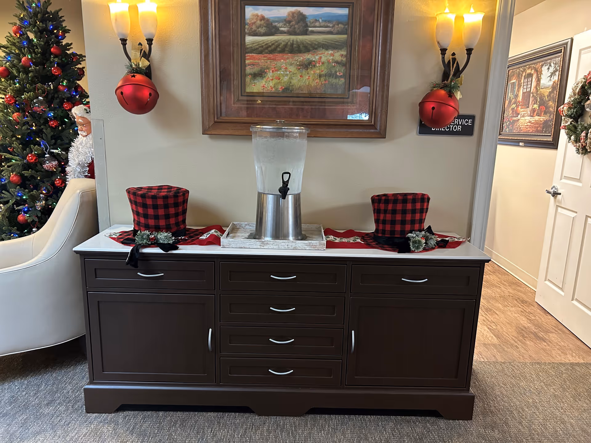 A dark wooden sideboard with a water dispenser on a white tray in the center. On either side of the dispenser are two red and black checkered top hats with small decorative greenery. Above the sideboard is a framed landscape painting. On the wall, there are two wall sconces with frosted glass shades and large red jingle bell decorations hanging from them. To the left, a decorated Christmas tree and a partial view of a white armchair are visible. To the right, a hallway with framed artwork and a door with a wreath is seen.