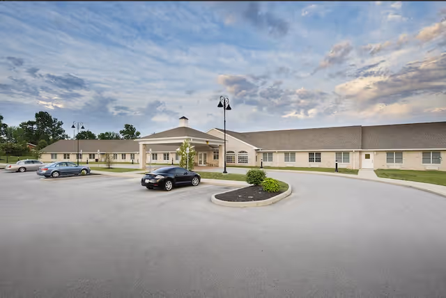 Front exterior of The Village Of Avon single-story senior living building with a circular driveway, parked cars, and a lamp post under a cloudy sky.