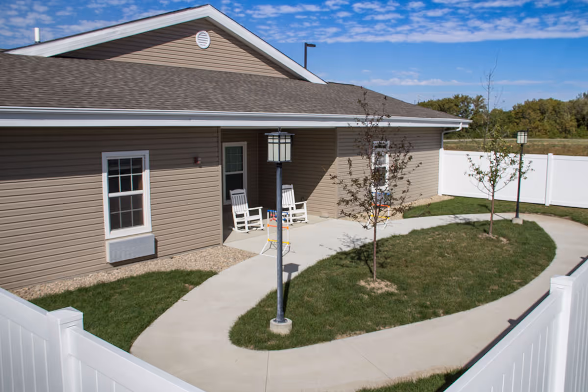Small enclosed courtyard with a curved concrete walkway, young trees, lamp posts, a white fence, and a beige building with a covered porch and rocking chairs.