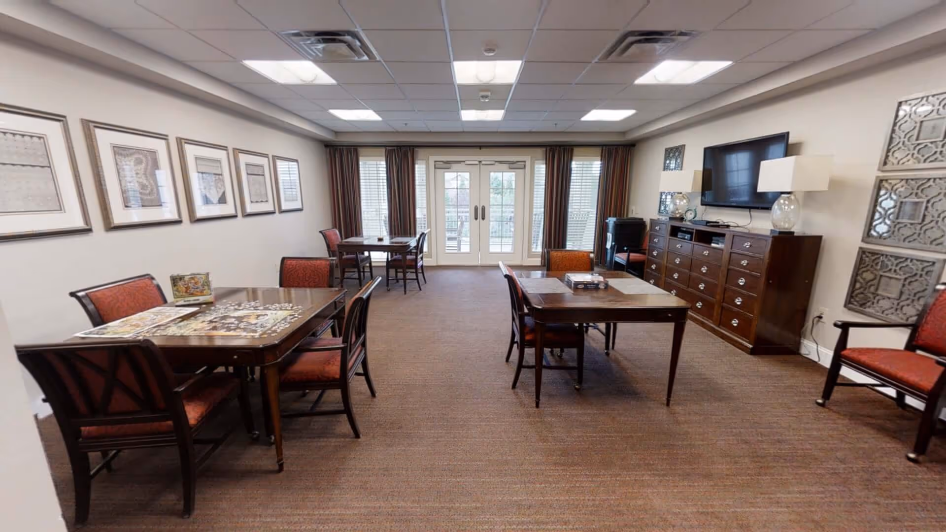 A spacious common room with several tables and chairs arranged for group activities or dining. The room features framed artwork on the left wall, a large cabinet with a flat-screen TV mounted above it on the right, and glass double doors with curtains at the far end leading outside. The room is well-lit with ceiling lights and has a carpeted floor.