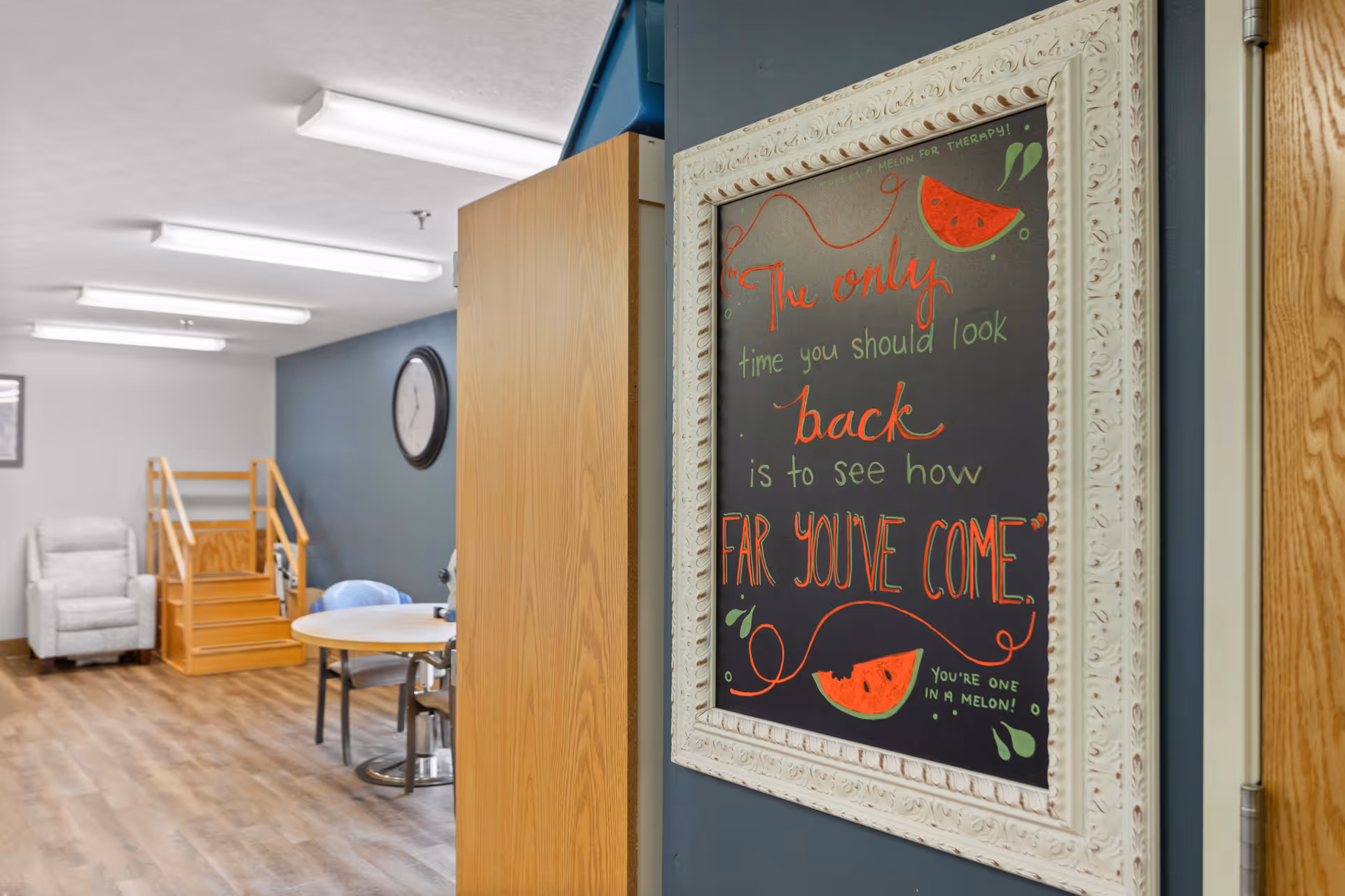 Interior view of a senior living facility showing a framed inspirational chalkboard sign on a wall with the quote 'The only time you should look back is to see how far you've come.' In the background, there is a small round table with chairs, a clock on the wall, a recliner chair, and a wooden staircase with handrails.