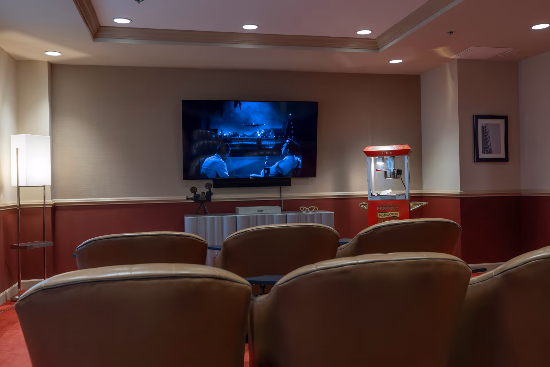 A cozy movie room with several leather chairs facing a wall-mounted flat-screen TV playing a black-and-white film. To the right of the TV is a red popcorn machine, and to the left is a tall floor lamp. The room has a warm ambiance with red and beige walls and recessed ceiling lights.
